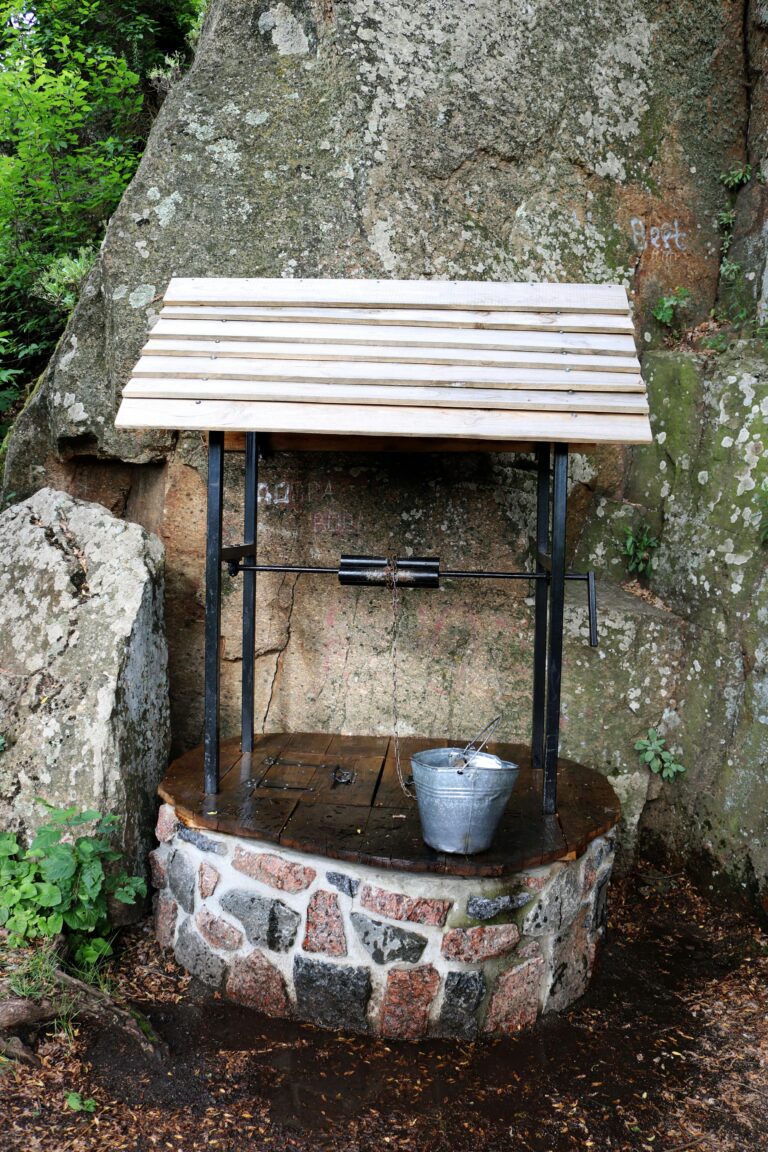 A rustic stone well with a metal bucket, set against a rocky backdrop in a rural setting.