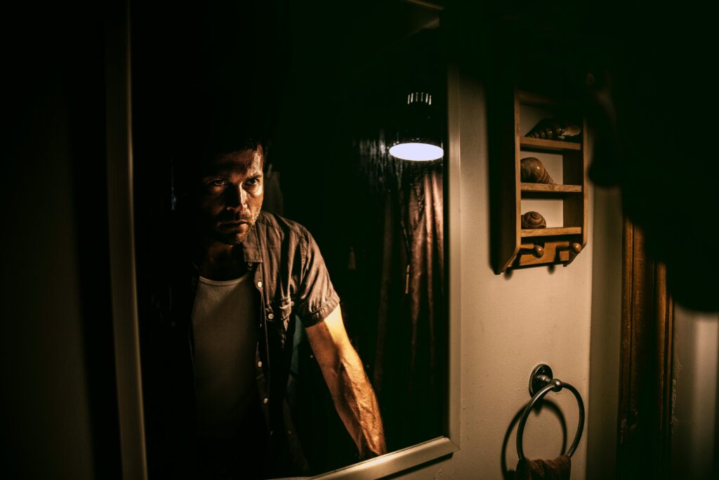 A man with a serious expression stands in front of a mirror in a dimly lit room.