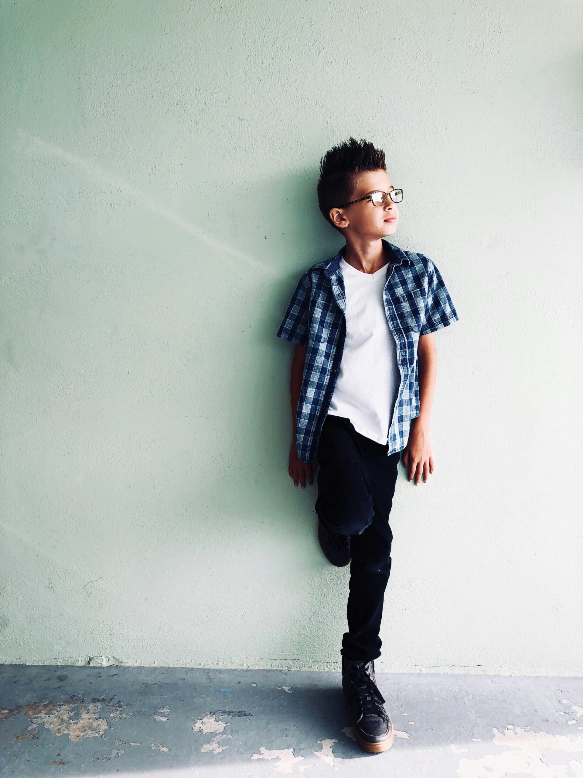 Fashionable young boy wearing plaid shirt, posing against a light wall, exuding stylish confidence.